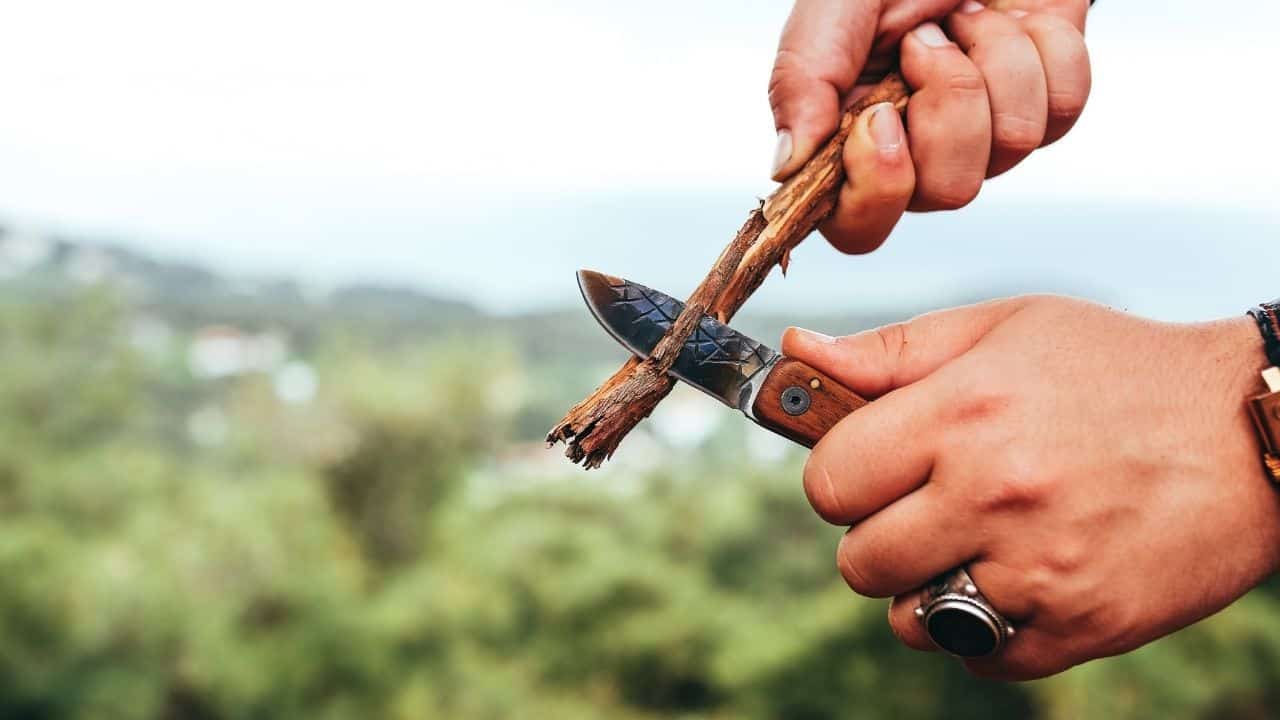 a photo of two hands using a folding knife to cut a piece of wood